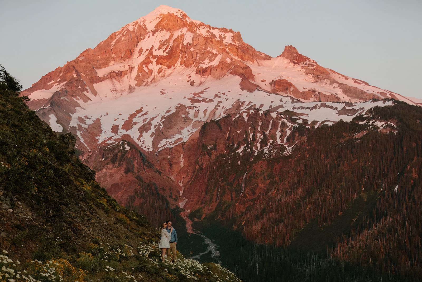 A couple stands at the base of Mount Hood during a sunset engagement photography session